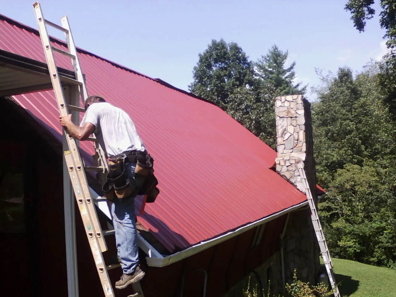 Roofer on a ladder beside a red metal roof and stone chimney — Soffit & Fascia Repair by Roofing Brothers in Peebles